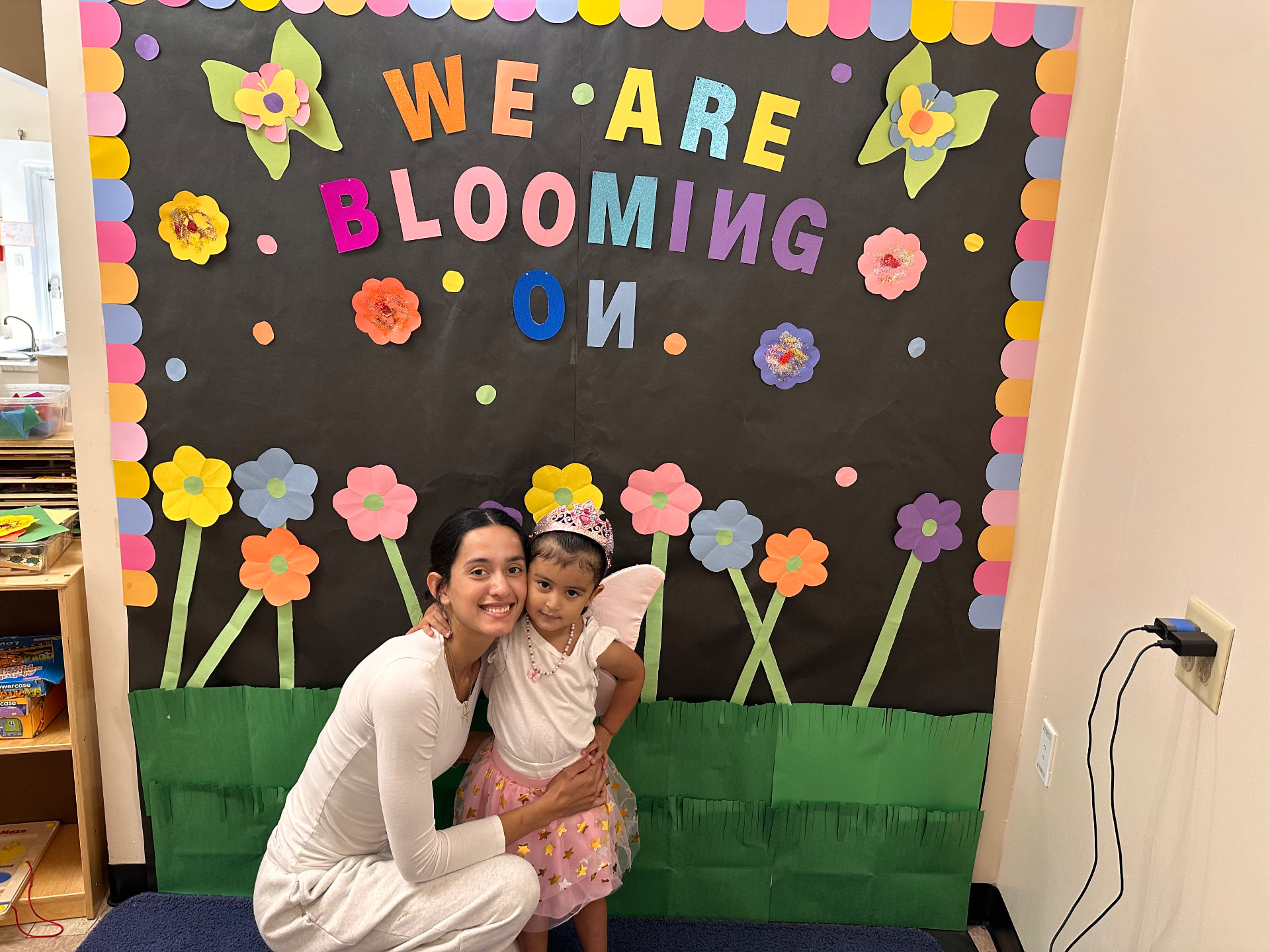 A mother and her daughter in front of a classroom bulletin board that reads We Are Blooming