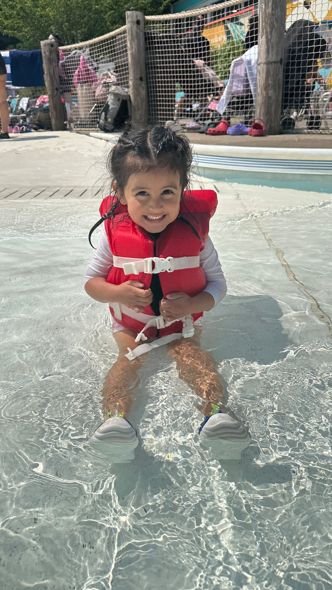 A little girl smiling in a red life jacket at the splash pad